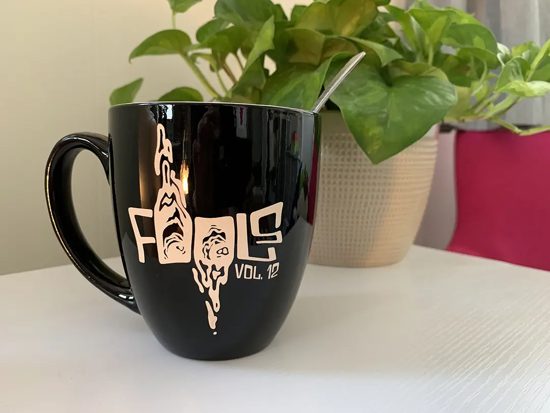 A black mug with a white graphic on it that says, 'Fools Vol. 12' in a distorted font. The Os in 'Fools' 
                        are manipulated to look like watery eyes. The left is dripping up, the right is dripping down.