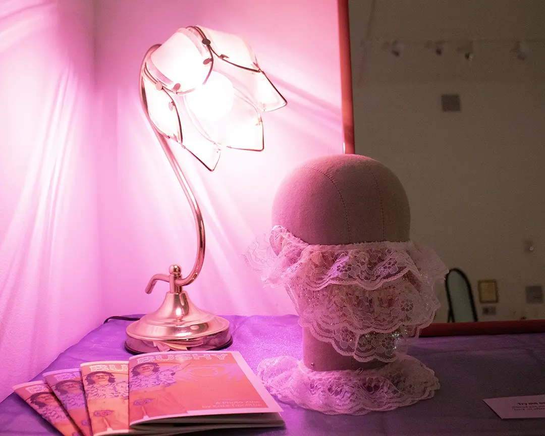 Mannequin head wearing a lace beard with a lace collar around the base. It's on 
                        a table with a sheer iridescent blue fabric over it, sitting in front of a mirror. 
                        Behind the head is a floral shaped lamp with a pink light casting patterns on the wall. 
                        There's also a stack of zines on the table.