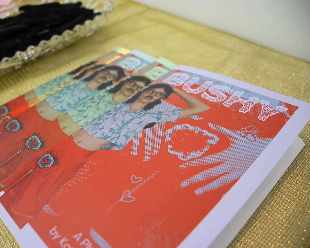 Close up of a stack of zines spread out on a pedestal. The pedestal is covered in a 
                        sheer, iridescent green fabric. Kate is on the cover of the zine wearing body hair clothes 
                        and accessories. The zine is titled the same as the show.