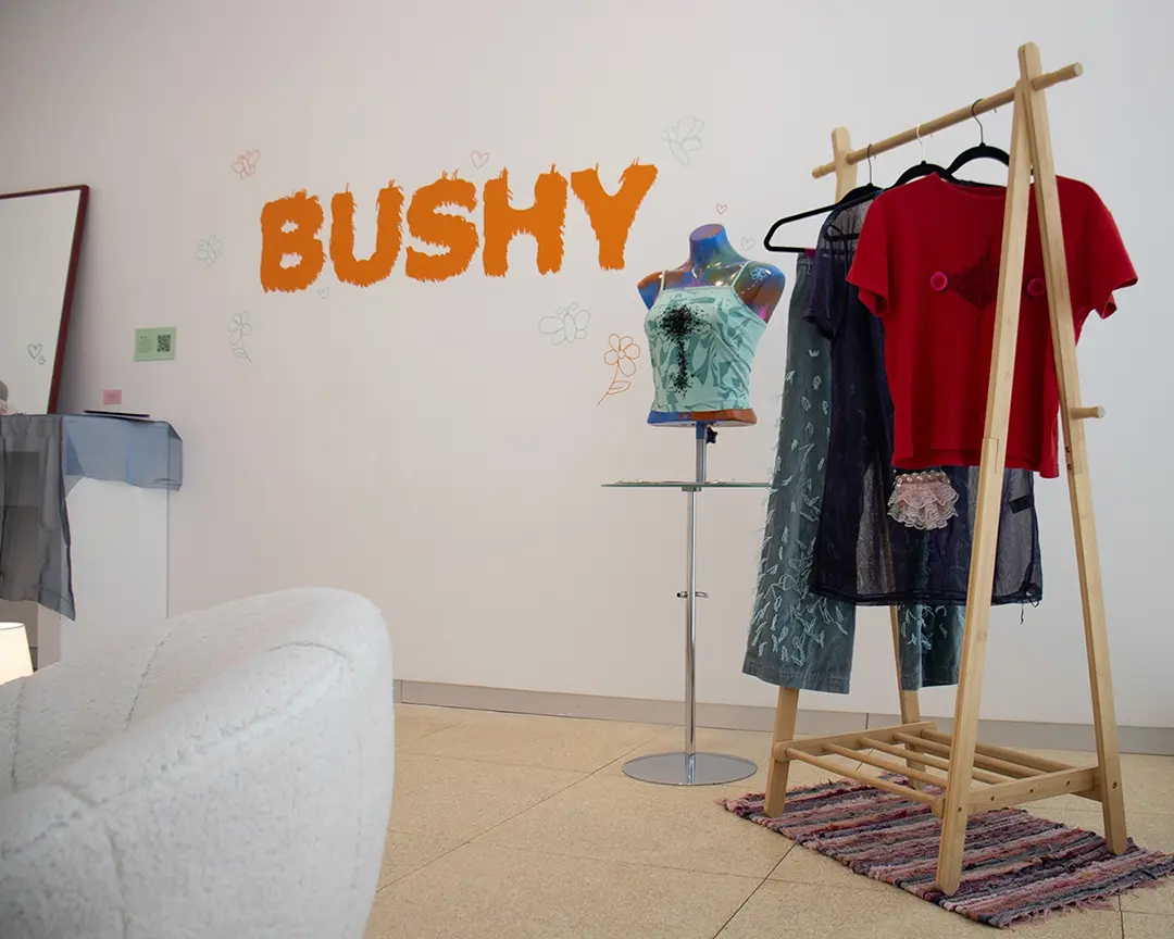 The back right corner of the gallery. There's a clothing rack with leg hair pants, 
                        a dress with a lace bush on it, and a shirt with chest hair and fake nipples on it. To the left 
                        of the rack is a small table with a mannequin bust on it wearing a black beaded chest hair shirt. 
                        On the wall is the title vinyl that says: BUSHY. There's a white fuzzy chair in the foreground 
                        partially off camera and a vanity setup in the back, also partially off camera.