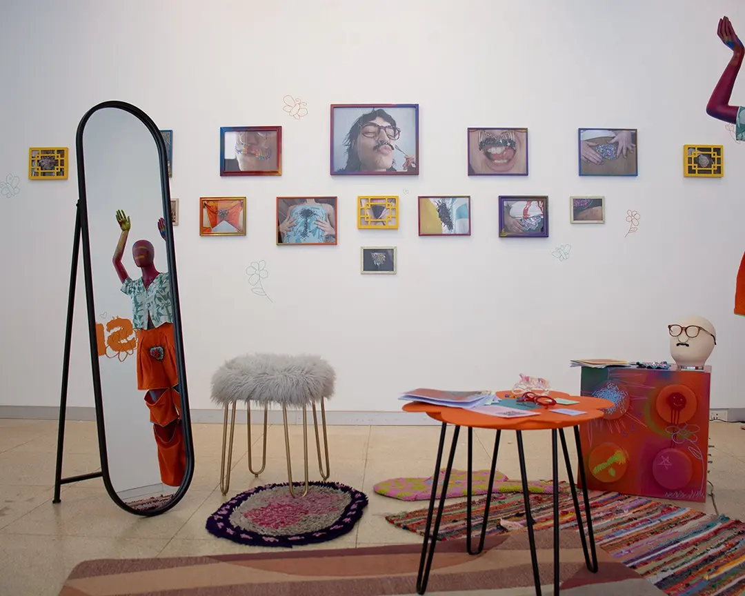 View of the left gallery wall which has many photos of people including Kate wearing 
                        body hair clothes and accessories. The photos are in colofrul frames of various sizes. In the 
                        foreground to the left is a floor-length standing mirror and a fuzzy stool. In the right-middle is an 
                        orange table shaped like a flower with zines and accessories on it. To the right is a table shaped like 
                        a giant lego with a mannequin head on it, and to the right of that, mostly off camera, is a full body 
                        mannequin with it's arm in the air.