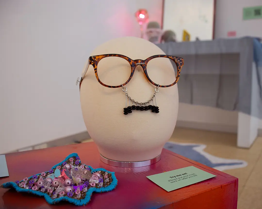 Close up of a mannequin head on a low table. It's wearing tortoise shell glasses frames with 
                        a black beaded mustache hanging from them. To the left is a pink, blue, and purple beaded bush in 
                        the shape of a butterfly made with butterfly shaped beads and sequins. Behind the head, out of focus, 
                        is a vanity set up.