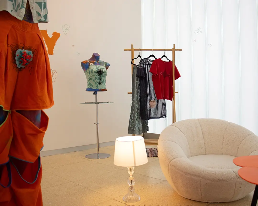 Another view of the back right corner of the gallery with a clothing rack and mannequin bust.
                        In the foreground to the left, partially off camera, is a full body mannequin wearing orange convertable 
                        pants with the zippers partially unzipped. In the foreground is a white fuzzy chair and a lamp giving 
                        warm soft lighting.