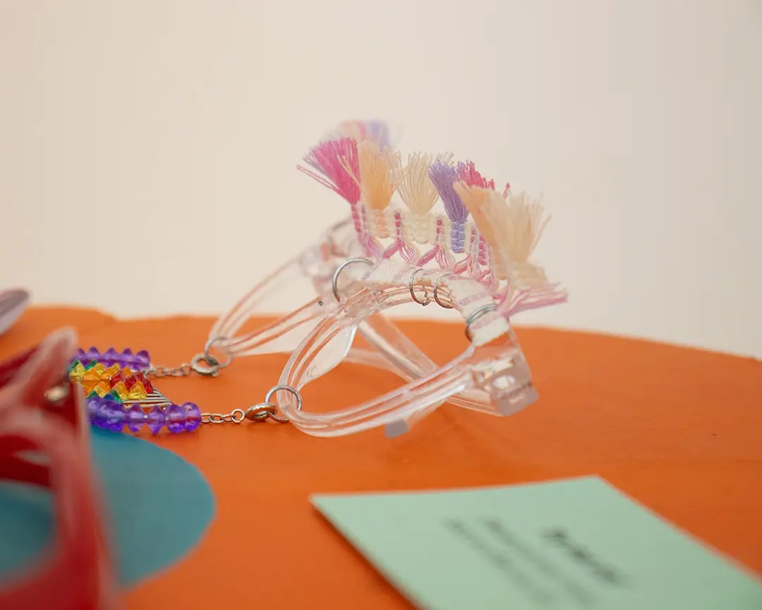 Close up of a pair of clear framed glasses on an orange flower table. They have a pink, purple, and yellow 
                        tassel trim attached to the top like a unibrow, and a rainbow beaded mustache hanging from the frames. There's 
                        a pair of red framed glasses out of focus in the foreground, slightly blocking the rainbow mustache.