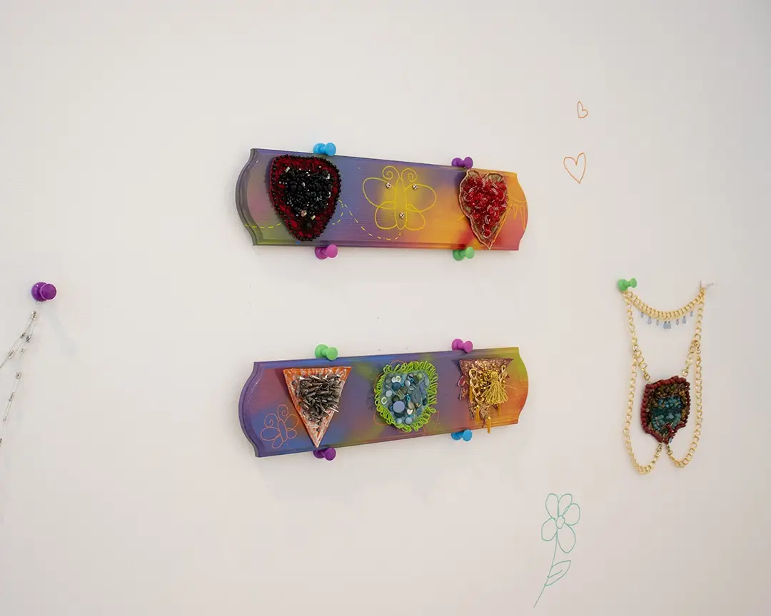 One wall of the gallery with two colorful boards hung up in the middle with large colorful 
                        push pins. On the top board are two mounted bushes with an empty space in the middle. 
                        The bottom board has three mounted bushes. To the left, out of frame, is a safety pin chain 
                        also hanging from a giant push pin. To the right is a harness, also hanging with giant push pins, 
                        that has a merkin in the shape of an archaic medusa head on it.