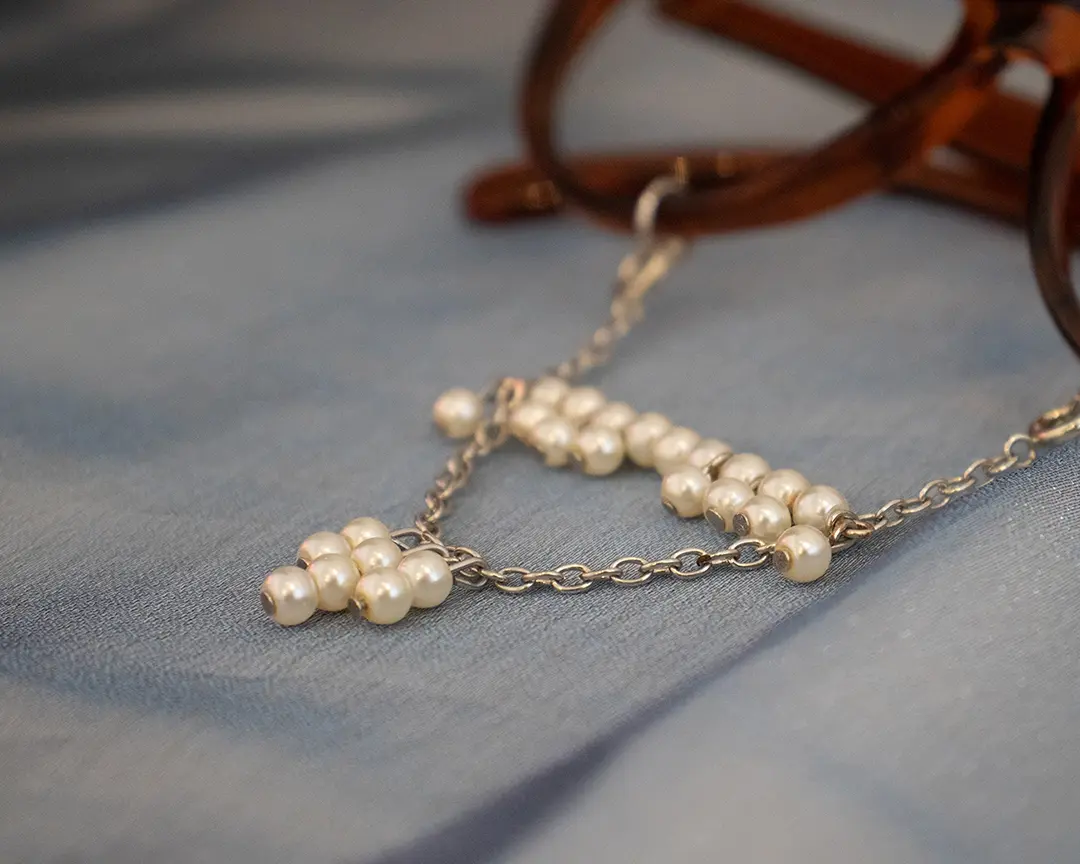 Close up of a pair of brown framed glasses with a beaded pearl mustache and soul patch 
                        hanging from the rims. It's sitting on blue iridescent fabric.