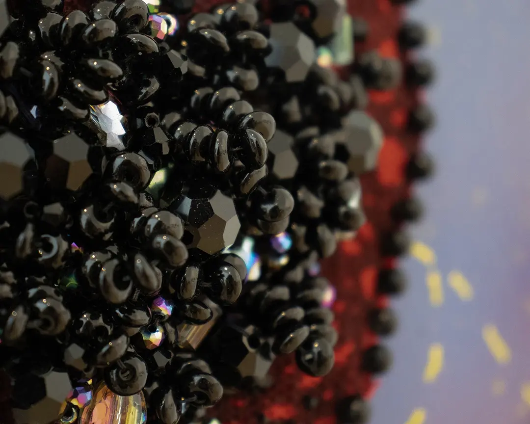 Close up of a beaded bush. It's made of smooth oval balck beads and iridescent 
                        beads forming a dense clump of pubic hair. It's sewn onto red fabric with a border of round 
                        black beads and it's mounted on a purple board.