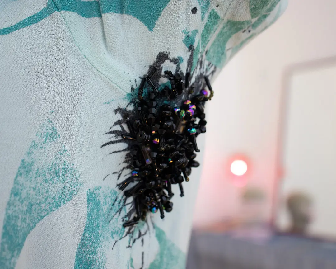 Close up of a shirt with beaded armpit hair on a mannequin. The black and iridescent beads 
                        are looped to look like thin strands of hair on the edges and more densly clustered hair in the 
                        middle. Out of focus in the background is a vanity set up.