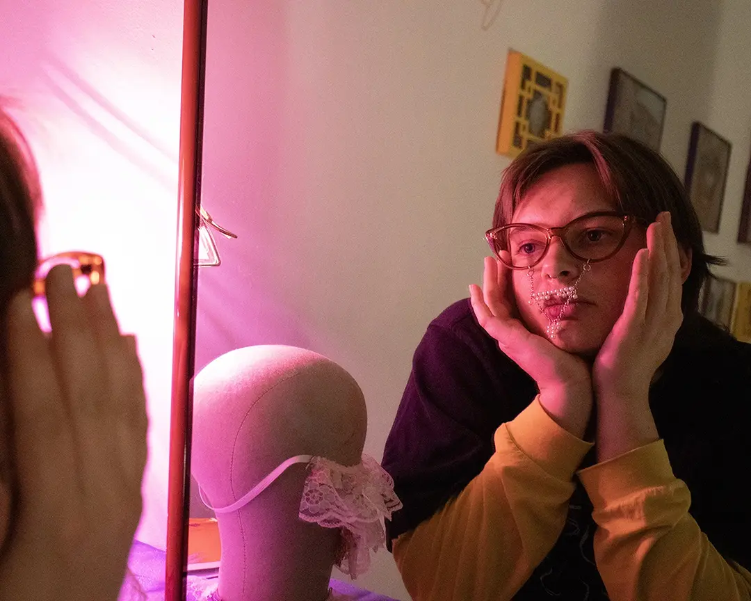 Nix looking in the mirror. The lighting is dark with a pink lamp casting a glow 
                        on their face. They're wearing brown framed glasses with a pearl mustache and soul patch 
                        hanging from them. Their chin is resting in their hands.