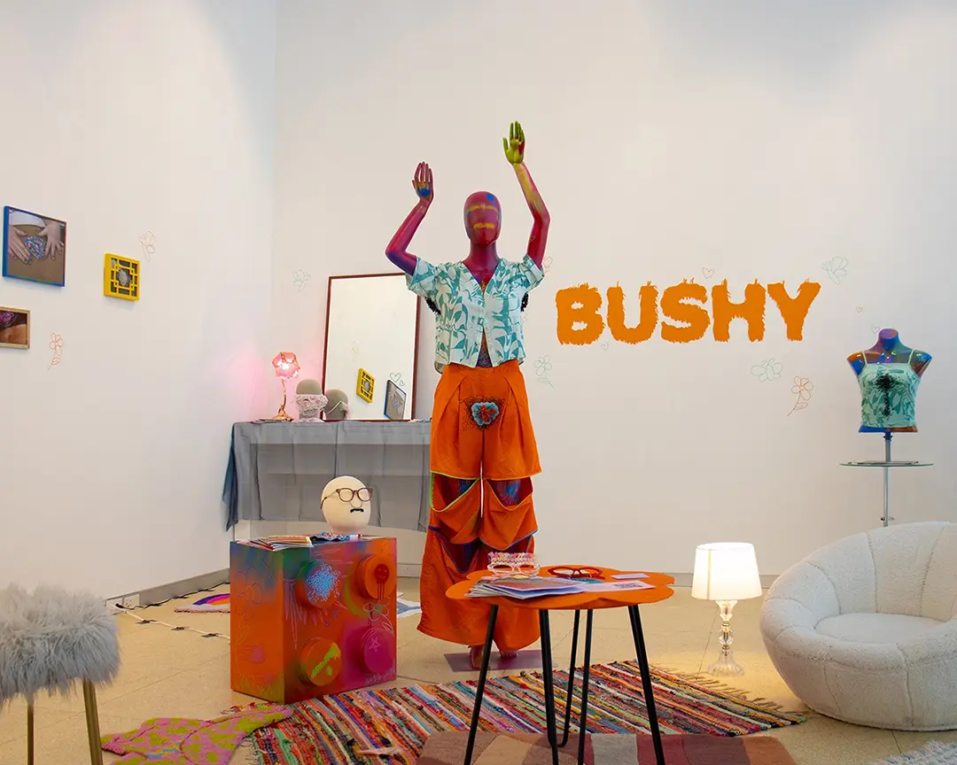 Wide view of a gallery with a pink mannequin in the middle. There are two 
                        small tables with various accessories around it as well as a fuzzy chair, a fuzzy 
                        stool, and a lamp. There are layered rugs on the floor. In the back is a vanity set 
                        up with a mirror, pink lamp, and accessories. On the other side is a table with a 
                        mannequin bust wearing a beaded chest hair shirt. On the back wall is hairy vinyl 
                        lettering that says the show title: BUSHY.