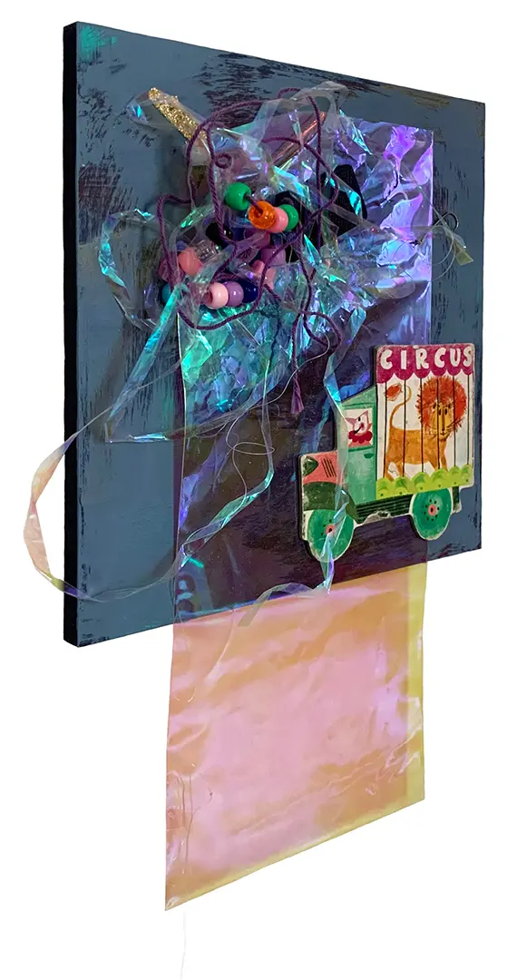 Left angle view of a three-dimensional collage on a painted board. It's made of miscellaneous 
                            trash, including strings of beads, plastic, and a circus cutout from a children's book.