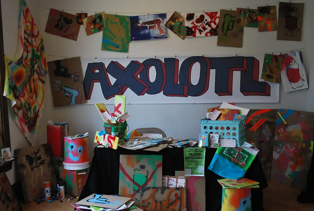 A table has spray painted cardboard signs, buckets, and wood panels, with zines, stickers,
                        and other materials scattered all around. The back wall has hanging cardboard signs and a large 
                        format sticker with the tag, 'AXOLOTL.'