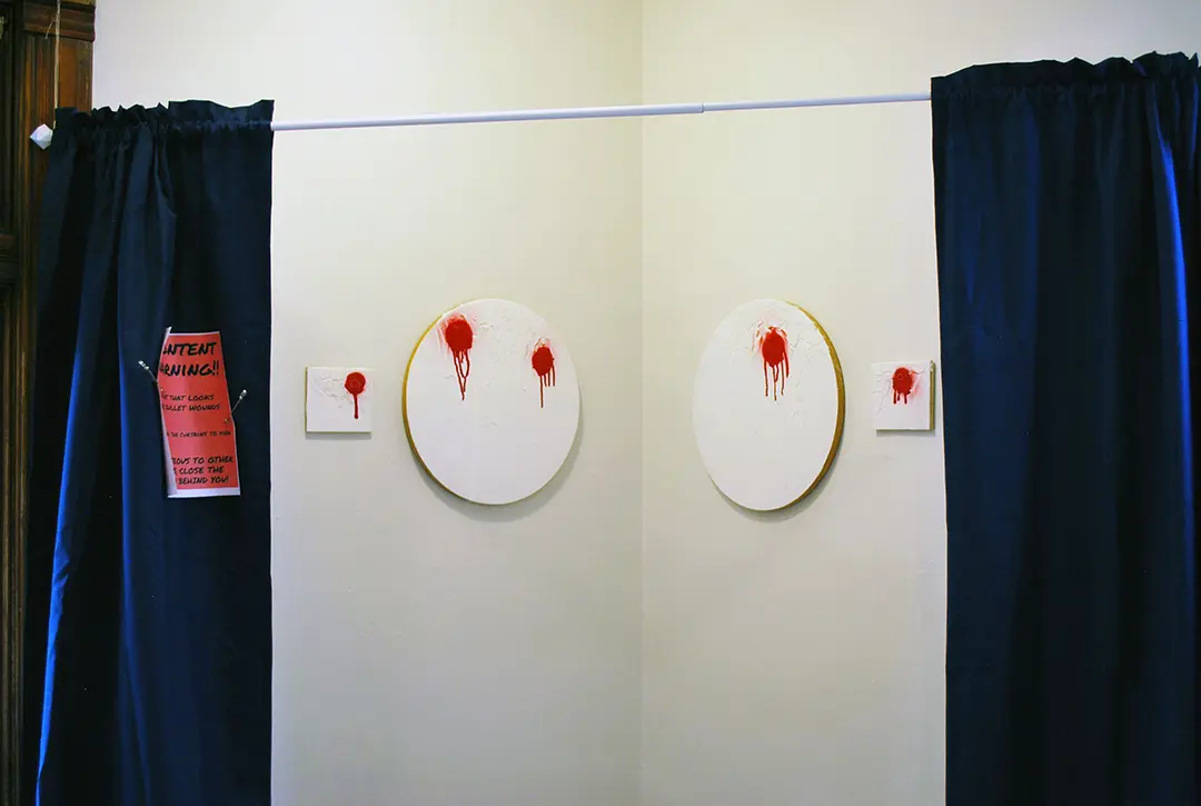 4 white paintings with red spray-paint that looks like bullet wounds. The paintings are behind 
                        a curtain with a content warning pinned to it, but the curtains have been pulled back for the photo.