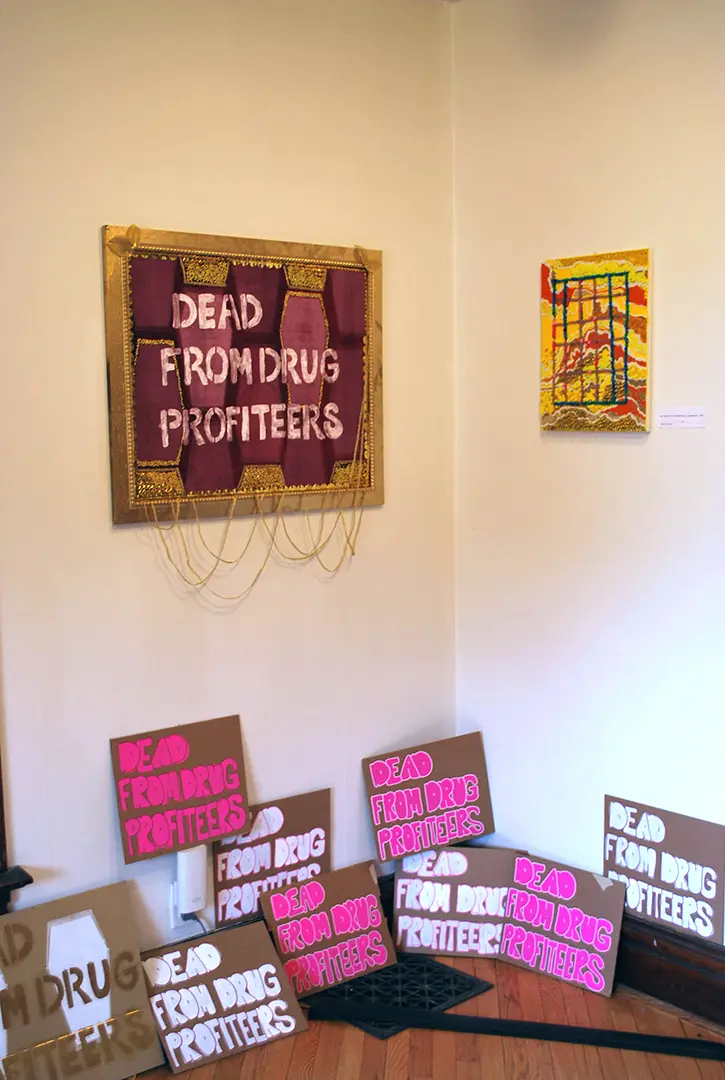 Velvet print in a gilded frame that says, 'DEAD FROM DRUG PROFITEERS.' 
                            Protest signs with the same phrase are on the ground. On the opposite wall is a bead painting.