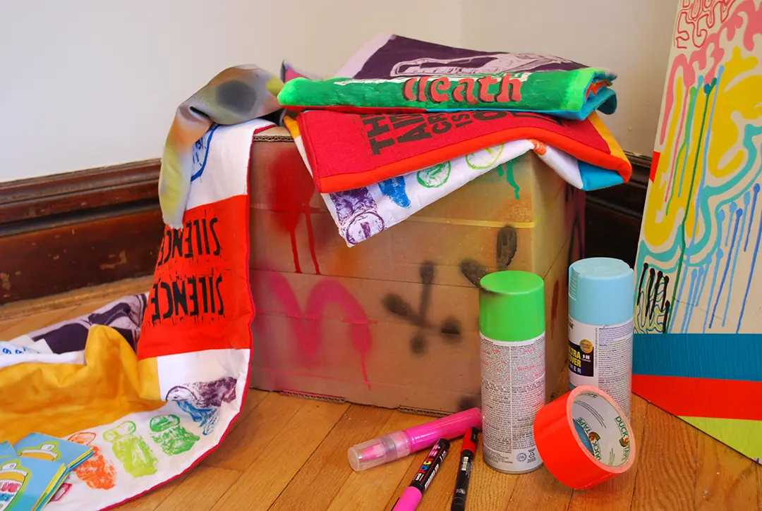 Close up of cardboard box and AIDS quilts. There are paint pens, spray paint, 
                        and duct tape on the ground.