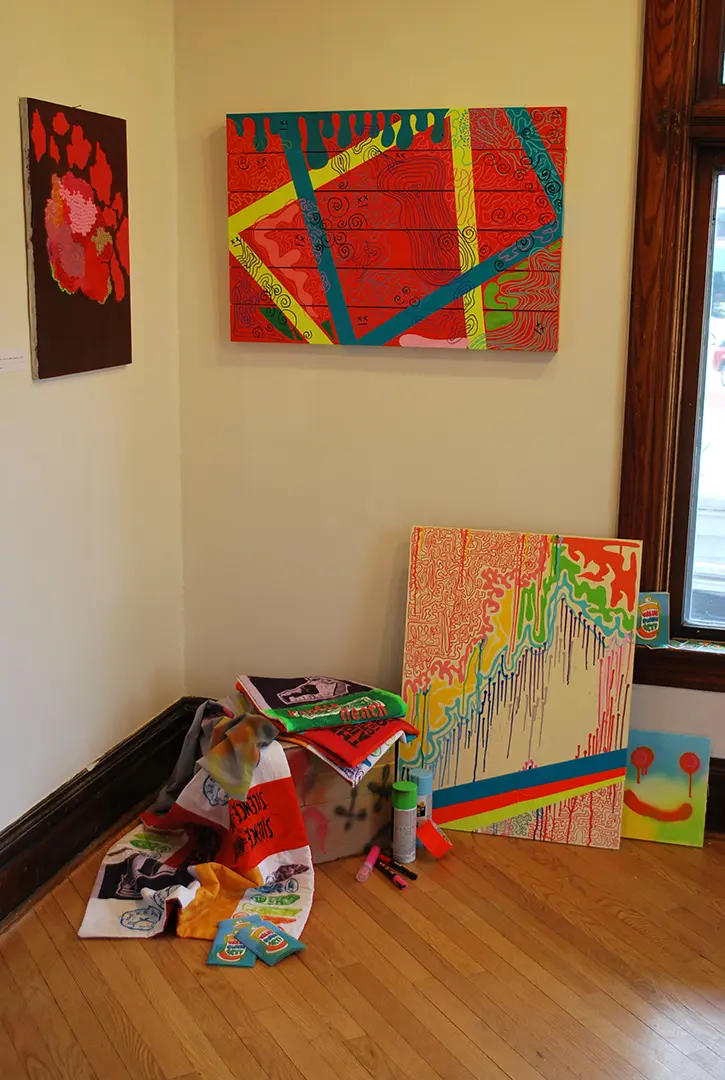 Bead painting on one wall and wooden painting on the other. On the floor is another 
                            wood painting, a cardboard box, and mini AIDS quilts.