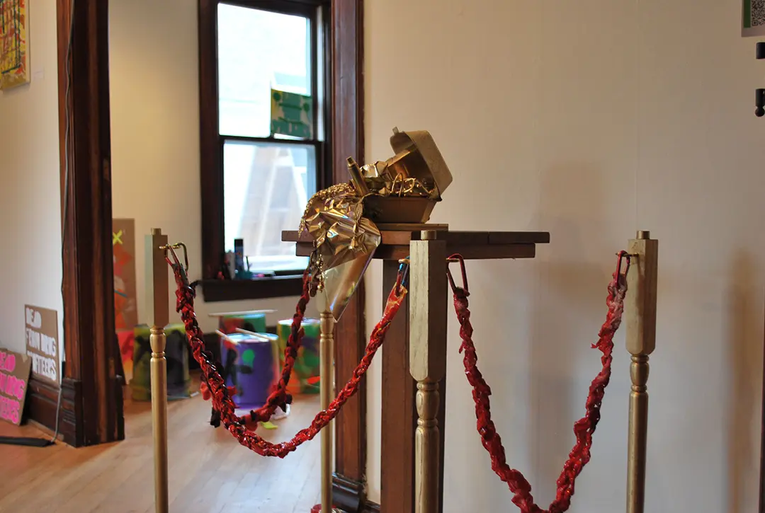 Wide angle of the treasure chest on the pedestal, surrounded by 'velvet' ropes made of trash 
                        and spray painted red. The ropes are clipped with carabiners to decorative wooden poles also spray painted gold.