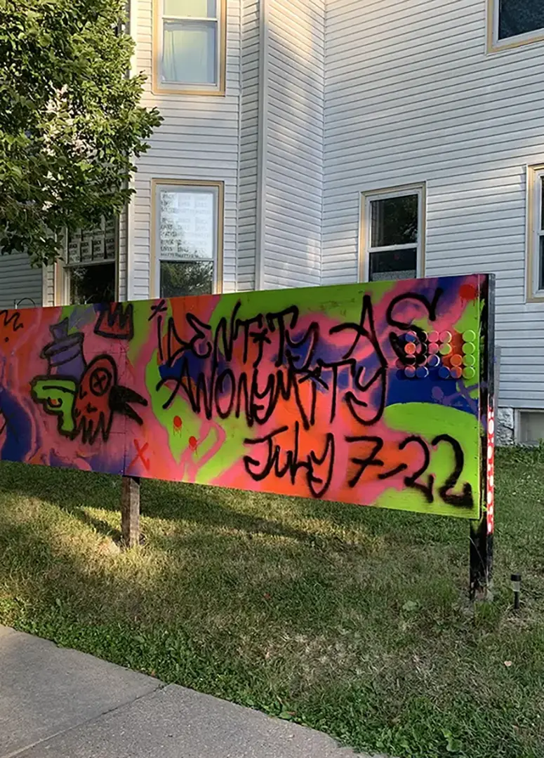 Long wooden sign spray painted with pink, blue, and green graffiti that says 'Identity as Anonymity, July 7-22.'