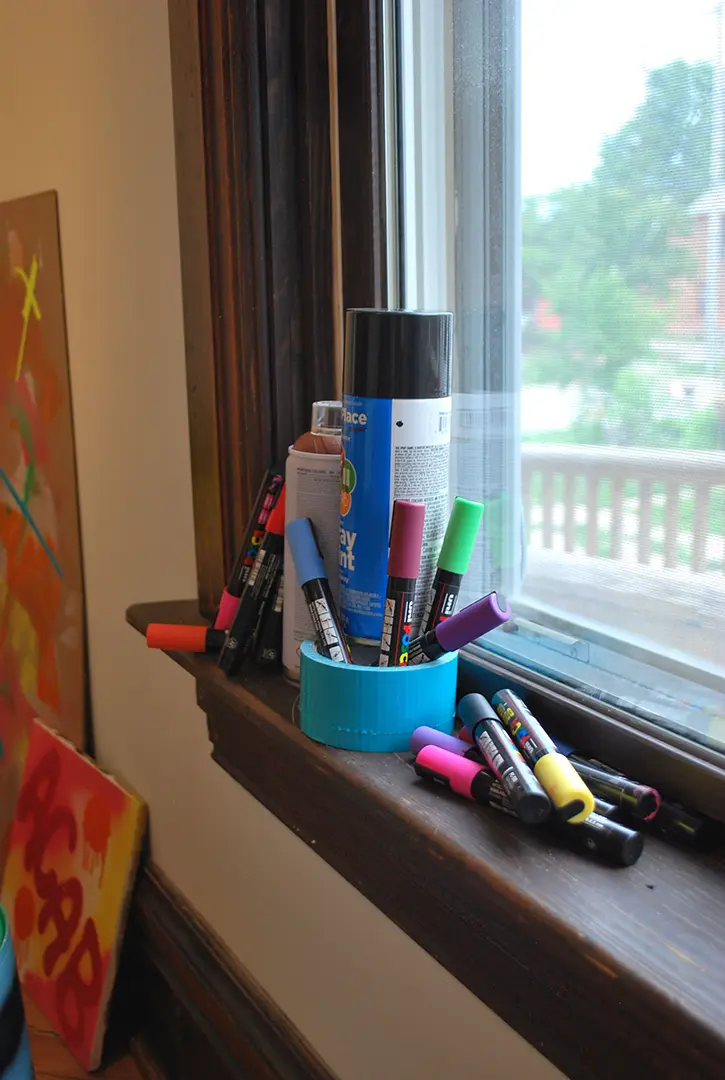 Spray paint cans, paint pens, and a roll of blue duct tape on a window sill.