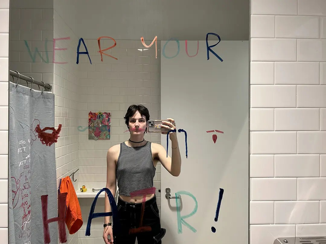 Mirror selfie of Kate. The mirror says "Wear your hair!" in expo markers and there are four facial 
                        hair variations for the viewer to stand in front of. Kate is standing in front of a pink mustache.