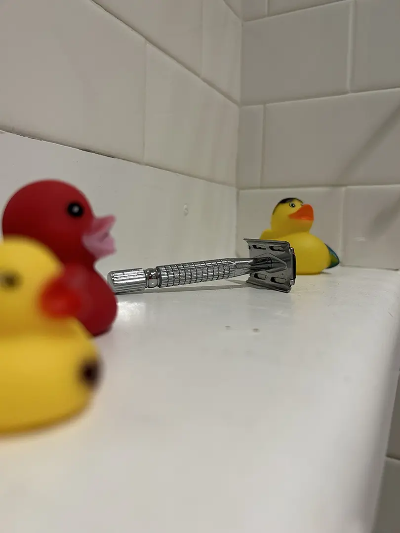 Close up of shelf with rubber ducks and safety razor. Two ducks are out of focus in the foreground, 
                            with the razor in focus in the middle.