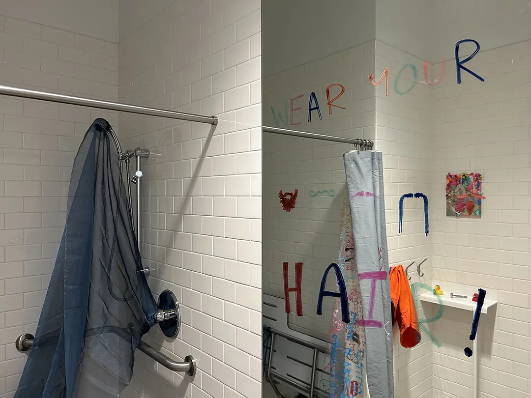 Close up of mirror and open shower. There are mustaches drawn on the mirror and text that reads: Wear your hair!
                        There's sheer blue fabric inside the shower.