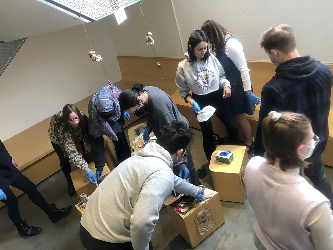 Still from a video of 'Sort Trash With Me!' Kate and their classmates are 
                        sorting trash that has been dumped on a pedestal in the Visual Arts Building.