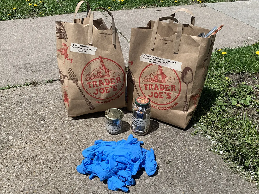 Two brown paper bags and two recycled jars full of categorized trash, 
                            and a pile of used blue nitrile gloves.