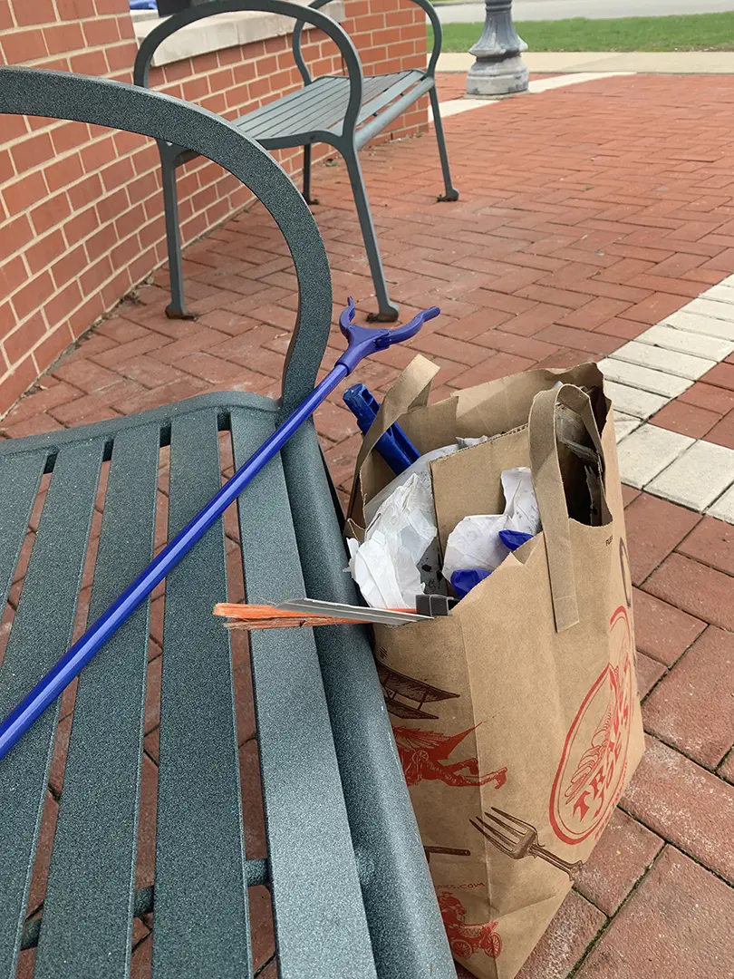 A brown paper bag filled with trash leaning against a bench. A blue reach 
                            extender is laying across the bench.