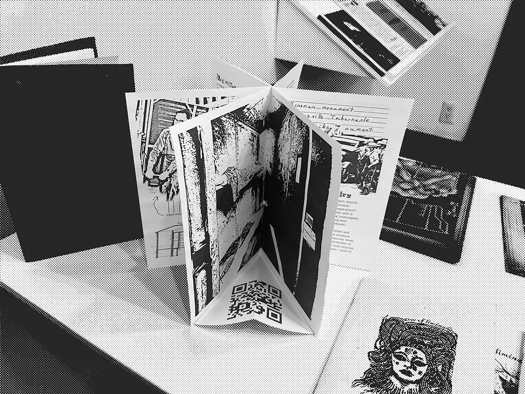 A zine standing on a pedestal. It's been bound rotary-style to emulate the 
                        jail, with each spread making up a pie wedge. The front facing spread is a full-spread photo of 
                        the inside of one of the cells, and has a paper floor with a QR code on it.