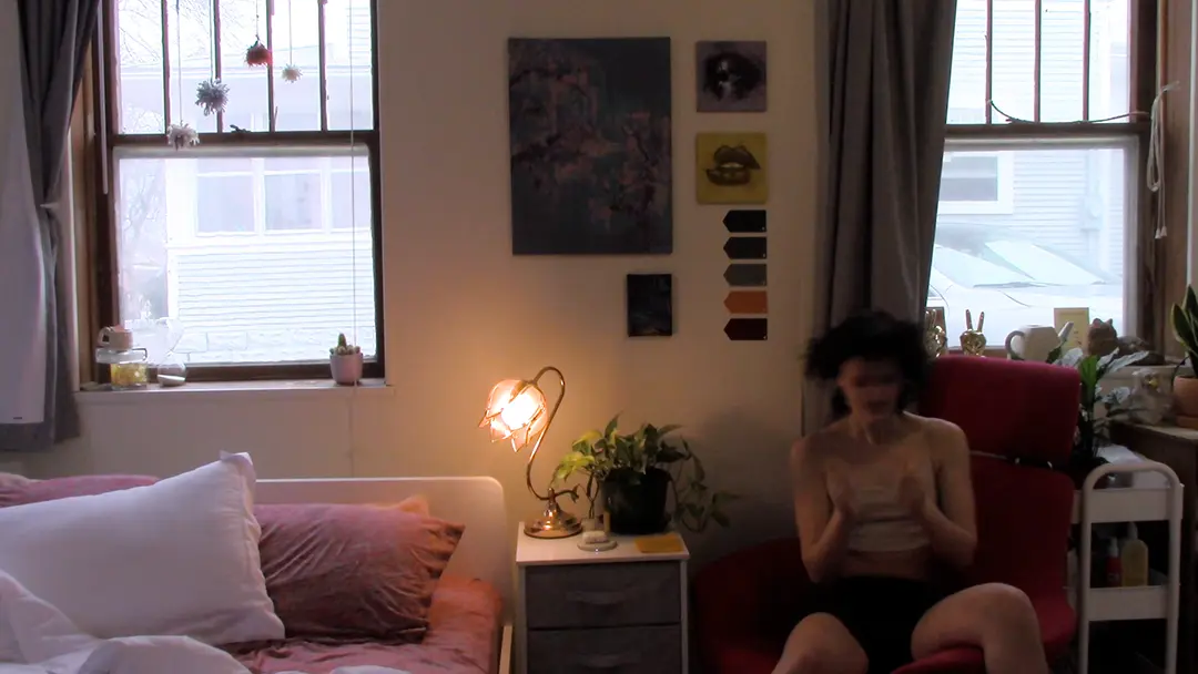 Wide still of Kate's room. She's sitting in a pink chair to the right side of the frame, air drumming.